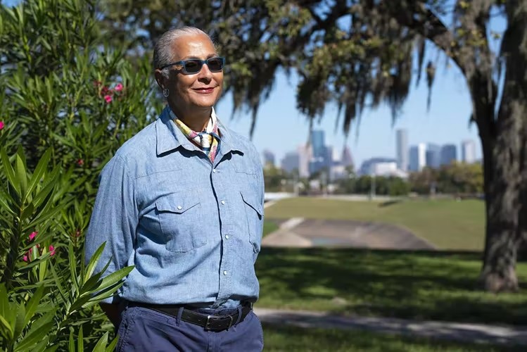 Elderly woman with sunglasses, standing at the side of a bush, with a view of the park in the background
